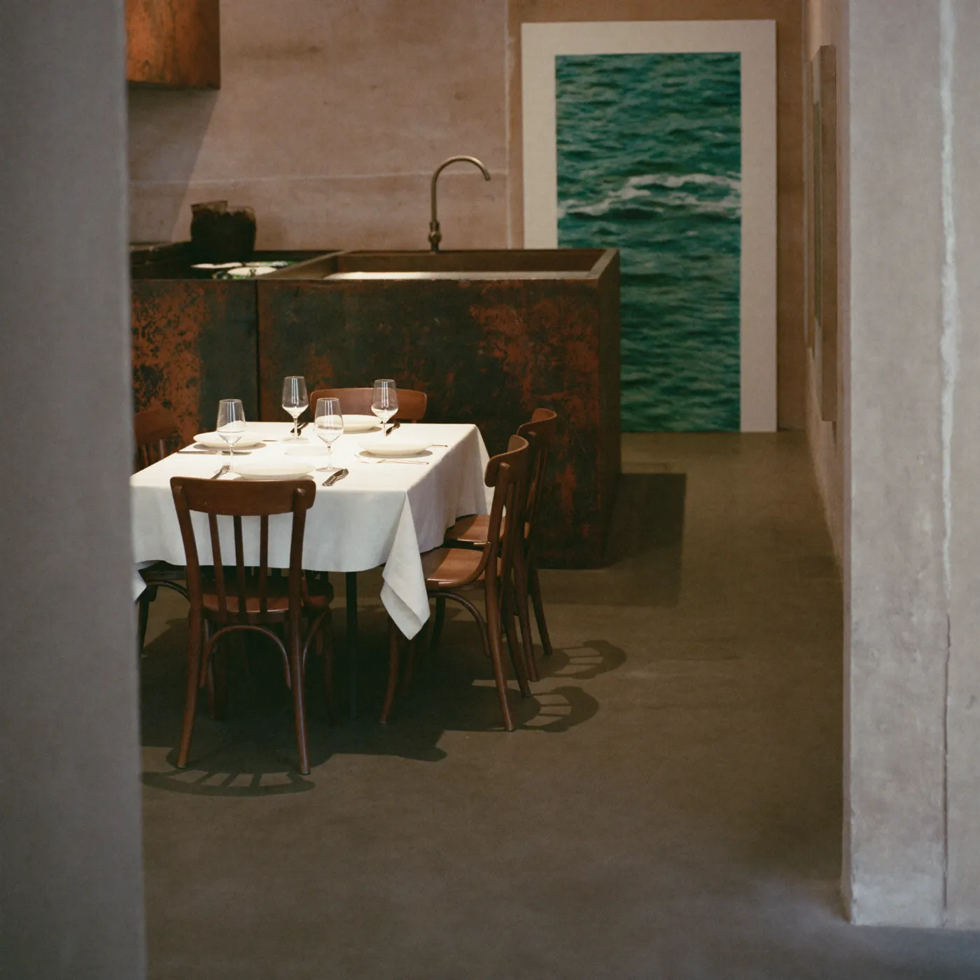 Salle de SILLON : ancien atelier réhabilité, bloc de cuisine en acier patiné, murs à l'enduit minéral, chaises en bois courbé et grande photographie de mer